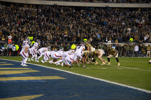 Fußballspieler in Sportkleidung und Helmen auf dem Boden während eines Spiels, mit Zuschauern und Stadionmerkmalen im Hintergrund.