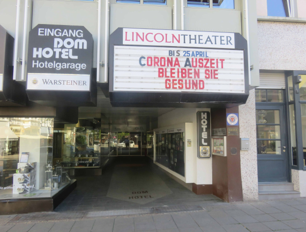 Außenansicht des Lincoln Theaters in Berlin, Deutschland, mit Glasfenstern und -türen und einer Tafel mit Text sowie einem Inneren, das eine pulsierende Stadtlandschaft suggeriert.