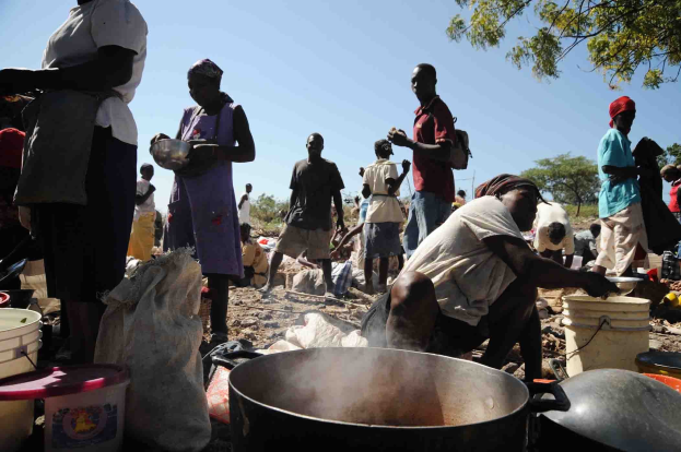 Eine Gruppe von Menschen bereitet im Freien Essen in einer ländlichen Gegend zu, umgeben von Eimern und anderen Gegenständen.