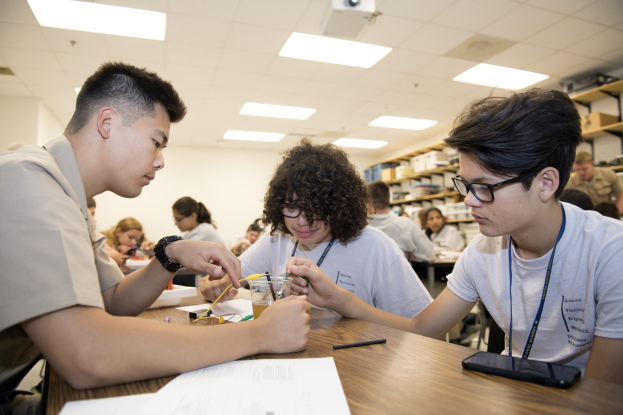 Three students at a classroom table working on a project with papers, pens, a mobile phone, and other objects; racks with items and ceiling-mounted lights and projector in the background.