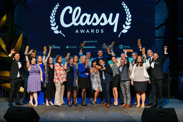 Eine Gruppe von Menschen auf einer Bühne, die lächeln und Auszeichnungen halten, mit Lautsprechern darunter und einem Bildschirm im Hintergrund, der "Classy Awards 2019" anzeigt.