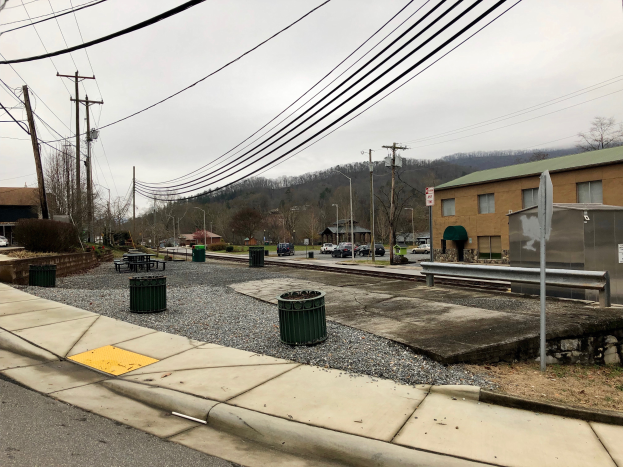 Eine Straßenszene in der Stadt mit Gebäuden, Fahrzeugen, Bäumen und einem bewölkten Himmel, mit einem Gehweg, Mülltonnen, einer Straßenlaterne, Strommasten mit Drähten und einem Müllcontainer.