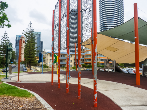 Ein Spielplatz in einer Stadt mit hohen Gebäuden im Hintergrund, umgeben von Gras, Bäumen und Pfählen, mit Fahrzeugen auf der Straße und einem bewölkten Himmel.