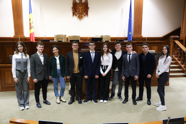 Gruppe von strahlenden Menschen, die vor einem Gerichtssaal mit Monitoren, Mikrofonen und anderen Gegenständen auf Tischen und einer hölzernen Plattform mit Flaggen und Stühlen im Hintergrund posieren.