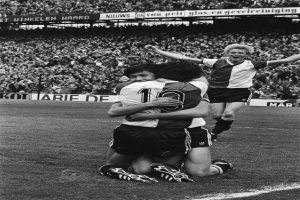 Drei Fussballspieler umarmen sich auf dem Boden und feiern ein Tor vor einer jubelnden Menge in einer schwarz-weißen Stadion-Szene.