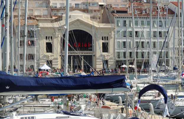 Ein Hafen mit zahlreichen Booten im Wasser, Gebäude mit Fenstern und einem Namensschild im Hintergrund sowie Zelte und Menschen.