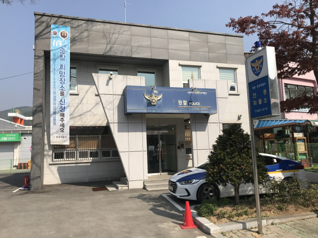 Ein Polizeiwagen steht vor dem Seoul Police Headquarters, einem Gebäude mit Glasfenstern und -türen, umgeben von Bäumen, Pflanzen und Verkehrskegeln, mit einer Fahne mit Text auf dem Gebäude und Hügeln und einem klaren blauen Himmel im Hintergrund.