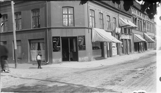 Ein Schwarz-Weiß-Foto eines Mannes, der eine belebte Stadtstraße mit Gebäuden, Pfählen und Geschäften entlanggeht.