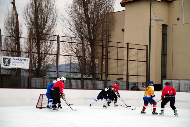 Menschen beim Eishockey auf einer Eisfläche mit Gebäuden, Bäumen, einer Straßenlaterne, einem Namensschild und Zäunen im Hintergrund unter einem klaren Himmel.