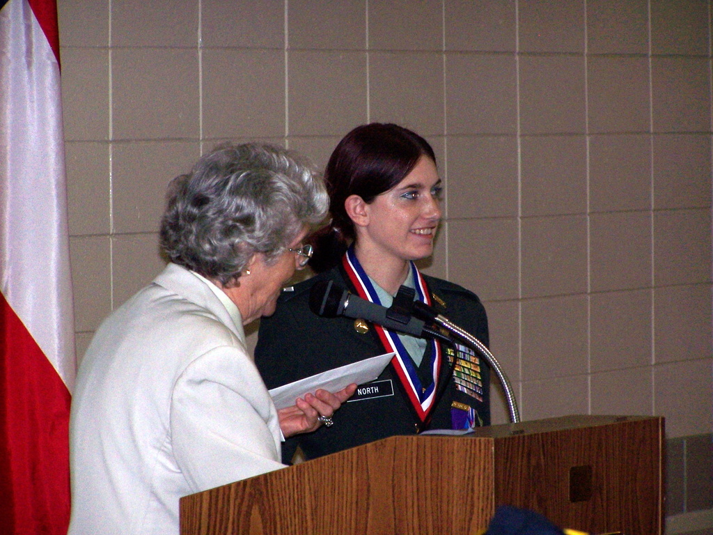 Zwei Frauen stehen vor einem Rednerpult mit einem Mikrofon, eine trägt eine Uniform, während eine Person links ein weißes Papier und eine Fahne hält.