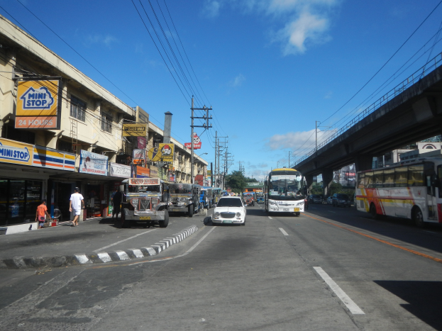 Eine belebte Straße in der Stadt mit Autos, Bussen, F"ußg"angern, Geb"auden, elektrischen Masten, einer Br"ucke, B"umen und einem bew"olkten Himmel.