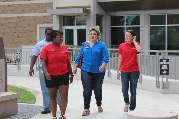 Vier Personen, drei Frauen und ein Mann, gehen einen Weg entlang mit schwarzen Tafeln mit Text an der Brüstung dahinter und einem Gebäude im Hintergrund.