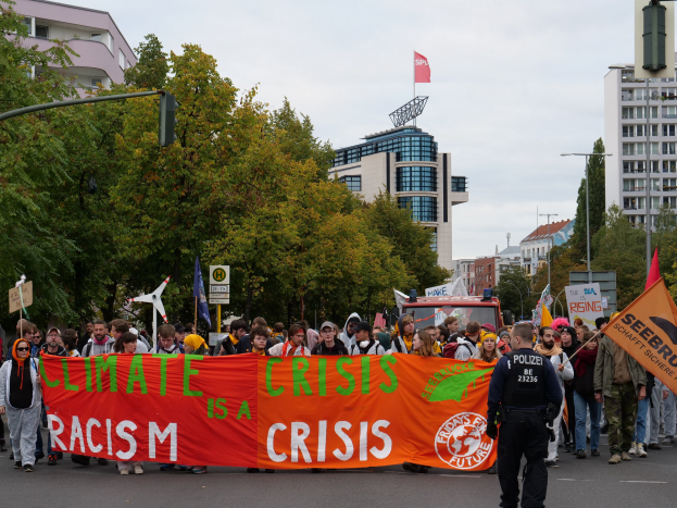 Eine Gruppe von Menschen marschiert eine von Bäumen gesäumte Straße entlang und hält ein Banner mit der Aufschrift "Klima-Krise ist eine Krise", mit parkenden Fahrzeugen, Schildern, Gebäuden und einem klaren blauen Himmel im Hintergrund.