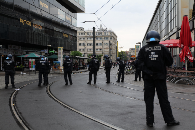 Gruppe von Polizisten in schwarzen Uniformen und Helmen auf einer von Gebäuden gesäumten Straße mit Glasfenstern, Bäumen, Polen, Drähten und einem klaren Himmel, mit verstreuten Schirmen, Stühlen und Tischen.