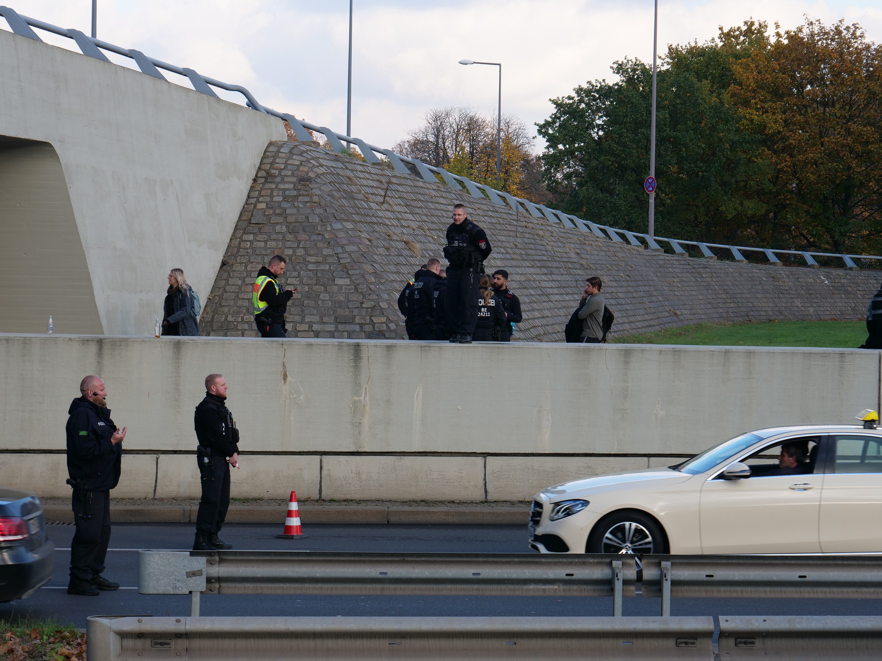 Eine Gruppe von Polizeibeamten steht neben einem Auto auf der Straße, mit Verkehrskegeln, einer Trennwand, Gras, einer Wand, Laternen, Bäumen und einem bewölkten Himmel im Hintergrund.