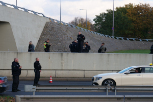 Eine Gruppe von Polizeibeamten steht neben einem Auto auf der Straße, mit Verkehrskegeln, einer Trennwand, Gras, einer Wand, Laternen, Bäumen und einem bewölkten Himmel im Hintergrund.