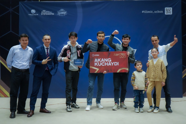 Gruppe von Menschen auf einer Bühne mit einer Tafel mit dem Text "Akang Kuchaydi - Zakovat Quiz", mit einem Banner, Barcode und Lichtern im Hintergrund.