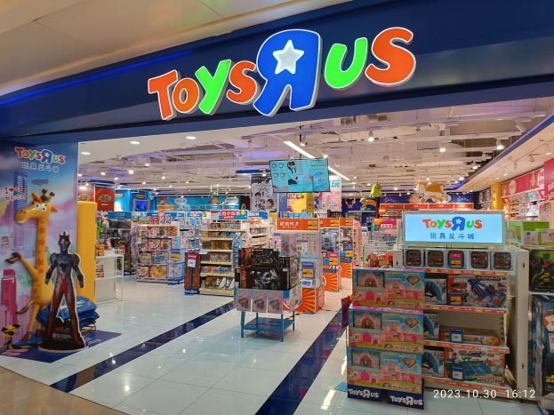 Ein Toys R Us Geschäft in einem Einkaufszentrum mit Regalen voller Spielzeuge, einem Namensschild mit Text, Deckenleuchten und einem Wasserzeichen in der rechten unteren Ecke.