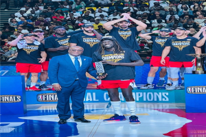 Männer mit Trophäen auf einem Basketballfeld mit Zuschauern im Hintergrund und Trophäen auf Podesten, die einen Sieg feiern.