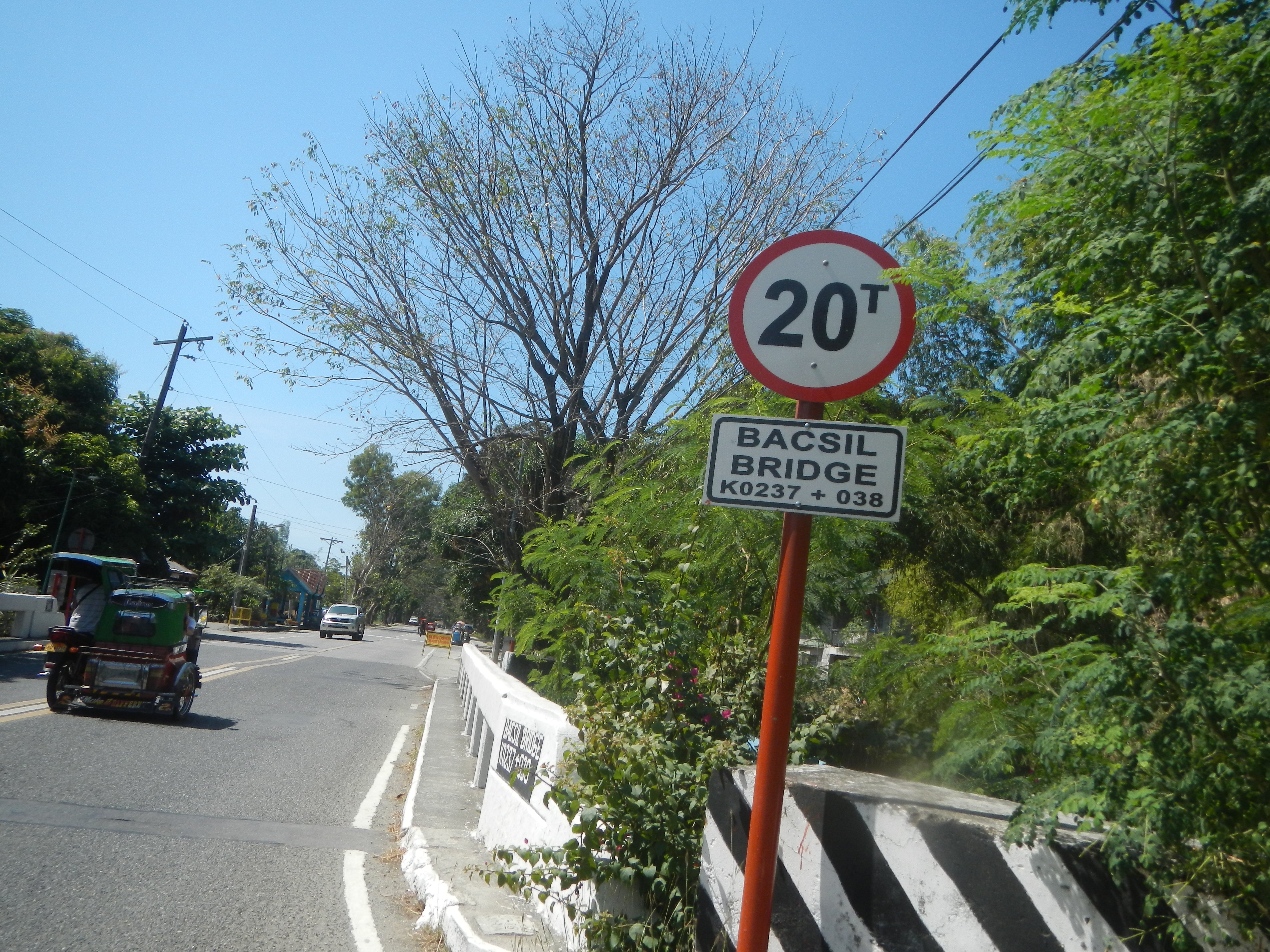 Eine Straße mit Fahrzeugen, ein Tempolimit-Schild, eine Trennlinie, Bäume, Strommasten mit Drähten und einen klaren blauen Himmel.