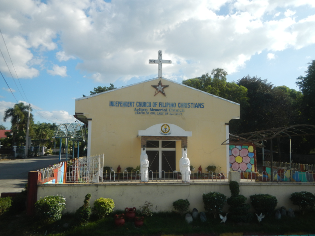 Eine unabhängige Kirche mit einem Kreuz davor, umgeben von einem Zaun, Statuen, Pflanzen, Gras, einem Weg, Bäumen, Pfählen, Drähten, einem nahen Haus und einem bewölkten Himmel.