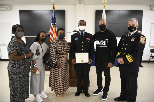 Eine Gruppe von Menschen mit Masken steht zusammen vor Flaggen, wobei die zentrale Person ein Zertifikat hält, in einer Umgebung, die an eine Abschlussfeier erinnert, mit Stühlen, Tischen und anderen Gegenständen im Hintergrund.