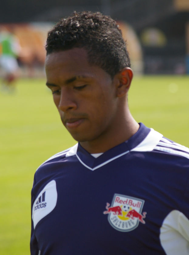 Ein junger Mann in einem blauen T-Shirt steht auf einem Fußballfeld, mit kurzen dunklen Haaren und einem entschlossenen Gesichtsausdruck, mit einem unscharfen Hintergrund.