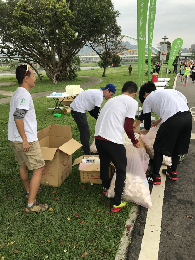 Gruppe von Menschen an einer Straße mit verstreuten Kartons und Flaschen, die an einem Charity-Lauf teilnehmen mit Bannern, Bäumen, Gebäuden und einem klaren blauen Himmel im Hintergrund.
