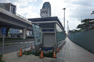 Buswartehäuschen am Straßenrand neben einem hohen Gebäude, umgeben von Verkehrskegeln, Pflanzen, einem Zaun, Fahrzeugen auf der Straße, Straßenlaternen, einer Brücke, Gebäuden und einem bewölkten Himmel.