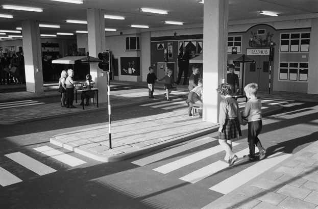 Schwarzes und weißes Foto von Kindern, die eine Straße an einer Fußgängerampel überqueren, mit Fußgängern in der Nähe, einem Verkehrssignalmast im Vordergrund und strukturellen Säulen mit Deckenleuchten im Hintergrund.