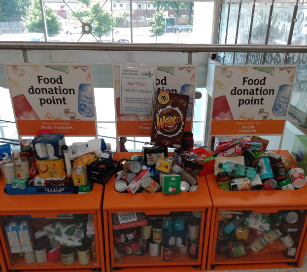 Orange Schübe mit Lebensmittelspenden, darunter Flaschen, Dosen und Kisten, mit Informationsschildern in der Nähe, durch Glasfenster mit Fahrzeugen, Bäumen, Gebäuden und Himmel im Hintergrund.