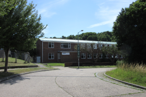 Ein Schulgebäude mit Fenstern, einem Straßenschild, einem Schild, einer Gruppe von Bäumen, Gras, einem Zaun und einem bewölkten Himmel, das in der Mitte eines Parks umgeben von saftigem Gras liegt.