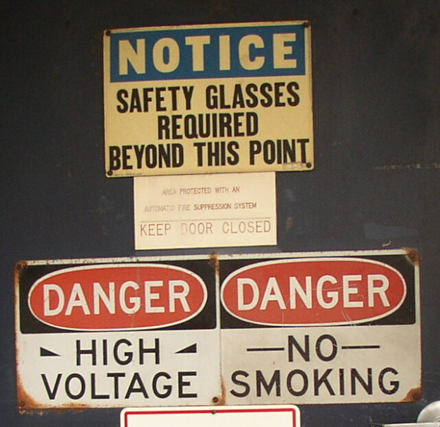 Ein Anschlagbrett mit drei leuchtend bunten Warnschildern, auf denen jeweils der Text "Notice: Safety Glasses Required Beyond This Point" in fetter Schrift steht.