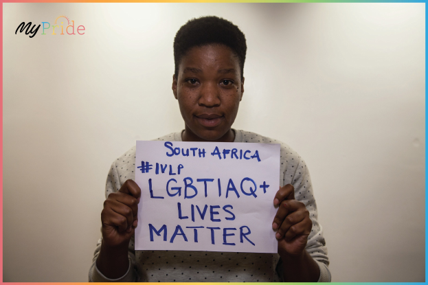Eine Frau in einem weißen Hemd steht entschlossen vor einer Wand und hält ein Schild hoch, auf dem in fetter schwarzer Schrift 'LGBTQ+ Lives Matter' steht.