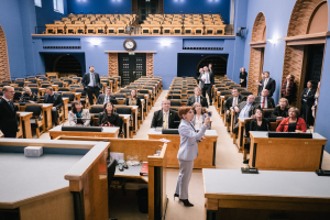 Eine Frau, die eine Rede im schottischen Parlament hält und vor einem Publikum steht, das auf Stühlen sitzt und steht. Auf den Tischen befinden sich Mikrofone, Gläser, Papiere und andere Gegenstände, eine Uhr an der Wand und Deckenleuchten im Hintergrund.