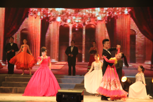 Eine Gruppe von Menschen auf einer Bühne mit Scheinwerfern, Stufen und einem Hintergrundbild, mit einem Lautsprecher unten, was eine Tanzvorstellung suggeriert.