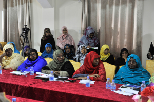 Eine Gruppe von Frauen sitzt an einem Tisch mit Flaschen, Büchern, Handys, Papieren, Stiften und einer Blumenvase, die in eine Diskussion vertieft sind, mit Vorh├Ąngen im Hintergrund.