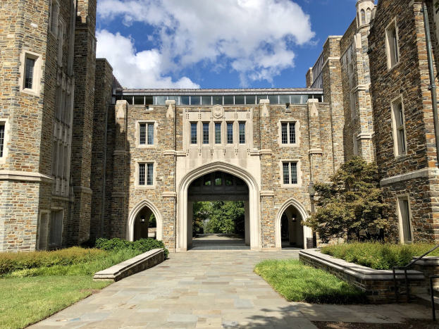 Ein großes steinernes Universitätsgebäude mit einem gewölbten Eingang, umgeben von grünem Gras, Pflanzen und Bäumen unter einem teilweise bewölkten Himmel.