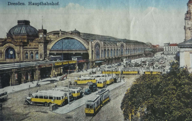 Ein sepiafarbenes altes Postkartenbild vom Dresdner Hauptbahnhof, das eine belebte Straßenszene mit Fahrzeugen, Fußgängern, Bäumen, Gebäuden und einem bewölkten Himmel zeigt.