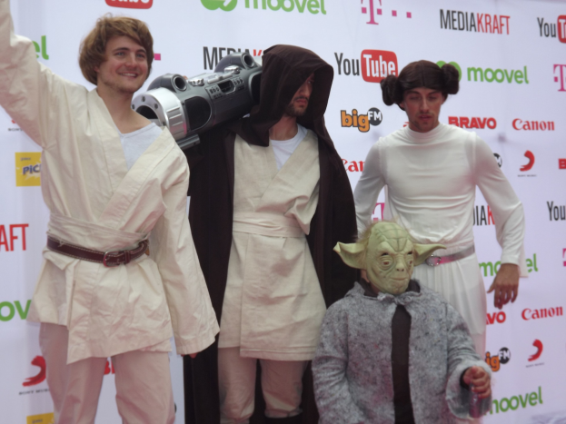 Eine Gruppe von Menschen in Star-Wars-Kostümen, die auf einem roten Teppich posen, wobei eine Person ein Objekt in der Mitte hält und ein Banner mit Text und Logos im Hintergrund zu sehen ist.