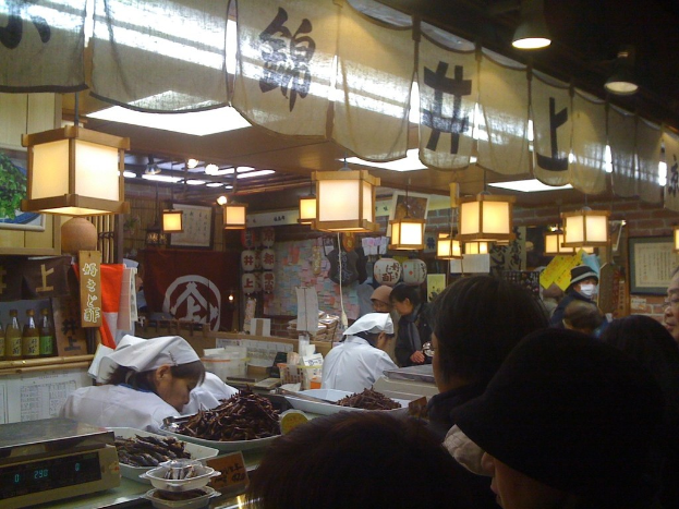 Restaurantmitarbeiter bereiten Artikel hinter einer Theke mit Kunden davor vor, während Flaschen mit Soßen, Menüs, Kleidung und Lampen im Hintergrund sichtbar sind.
