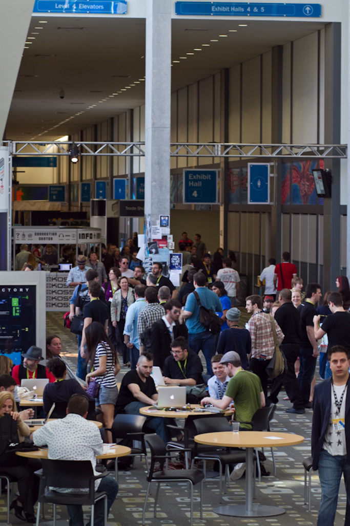 Ein belebter Flughafenempfang mit wartenden Menschen, einige stehen und andere arbeiten, Richtungsschilder an der rechten Wand und darüber sowie von der Decke hängende Lampen.