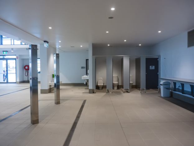 Große öffentliche Toilette mit mehreren Toiletten, tragenden Säulen, einem Mülleimer, einer Tür, einer Tafel, einem angebrachten Feuerlöscher und Deckenleuchten unter einem Dach.