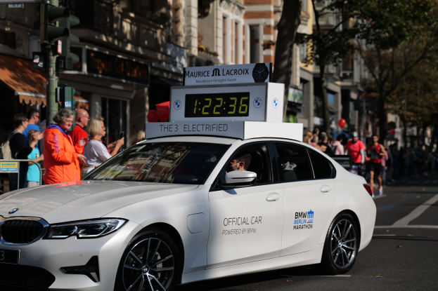 Ein weißer BMW mit einer Uhr auf dem Dach fährt eine Straße entlang, mit einer Person darin und einer Gruppe von Menschen auf der Straße, mit Gebäuden, Bäumen und Verkehrszeichen im Hintergrund.