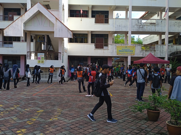 Eine Gruppe von Menschen, einige mit Taschen, geht vor einem Gebäude mit Fenstern, Geländern, Säulen, Bannern, Flaggen, einem Zelt, Topfpflanzen und Bäumen unter einem bewölkten Himmel.