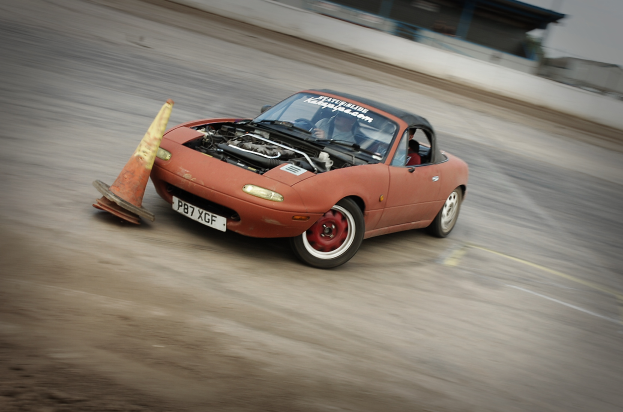 Eine Person fährt ein rotes Auto auf einer Rennstrecke, überholt einen Verkehrskegel mit einem unscharfen Hintergrund, der hohe Geschwindigkeit andeutet.