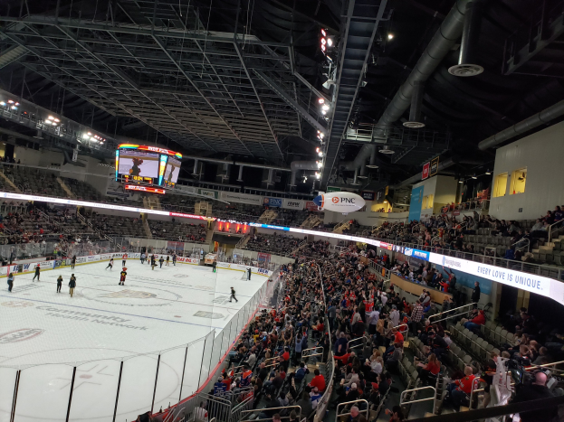 Eishockeyspiel in einer großen Arena mit sitzenden und stehenden Zuschauern in der Nähe der Eisfläche, umgeben von Bannern, Fenstern und Deckenbeleuchtung.