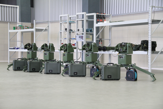 Eine Gruppe von Militärrobotern ist auf einem Boden positioniert, umgeben von Koffern und anderen Gegenständen auf Regalen, mit einer Wand und einem Geländer im Hintergrund.