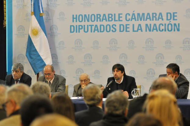Eine Gruppe von Menschen sitzt an einem Tisch mit einem Krug, Papieren und einem Stift, während eine Person in der Mitte in ein Mikrofon spricht, mit einer weißen und blauen Flagge und einem Hintergrund, auf dem "ehrenwerte kamara de" steht.
