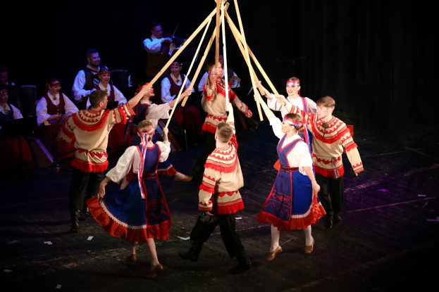 Eine Gruppe von Menschen in bunten Kostümen tanzt um einen Maibaum, einige halten hölzerne Stöcke, während Musiker im Hintergrund Instrumente spielen.